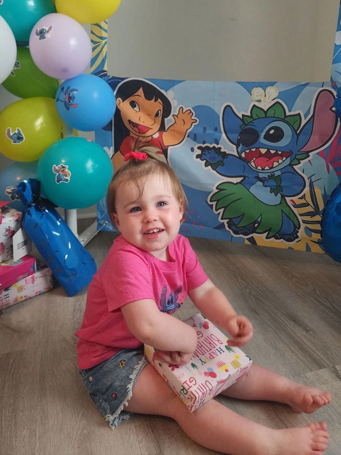 A young girl sitting on the floor holding a wrapped gift at a birthday party with Stitch and Lilo from Lilo & Stitch and balloons in the background.
