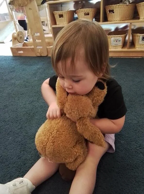 A young child sitting on a dark carpeted floor, hugging a plush brown teddy bear. The child has light brown hair and is wearing a black shirt and light-colored shorts. In the background, there are shelves with baskets and a wooden play structure.