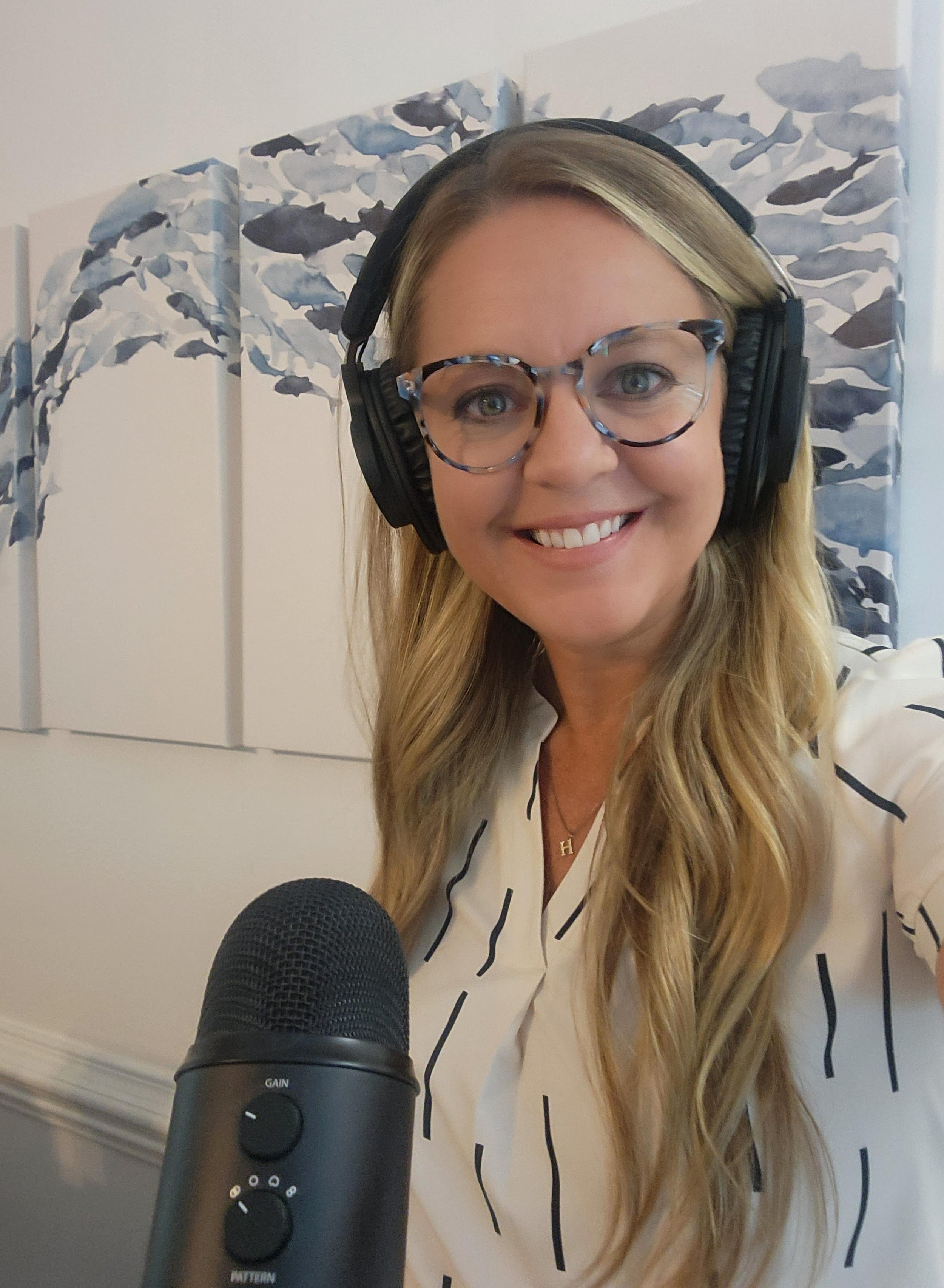 A smiling woman with long blonde hair, wearing glasses and headphones, sitting in front of a microphone, with blue and gray whale artwork in the background.