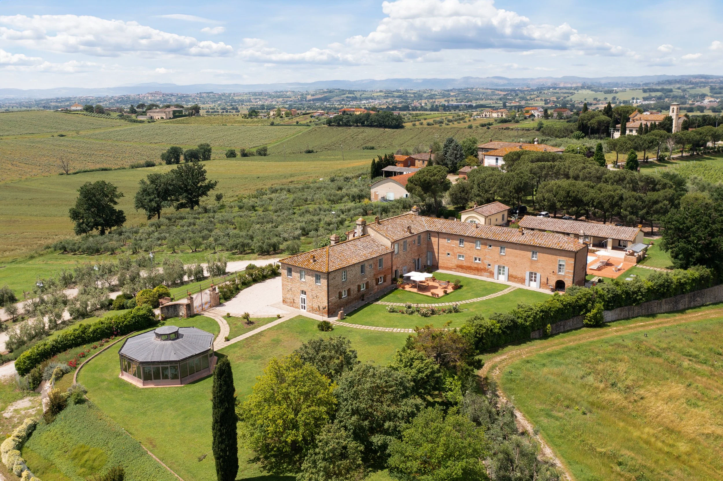 An aerial view of a large estate with a main brick house, a separate gazebo structure, and extensive landscaped gardens, surrounded by rolling fields and farmland in the countryside.