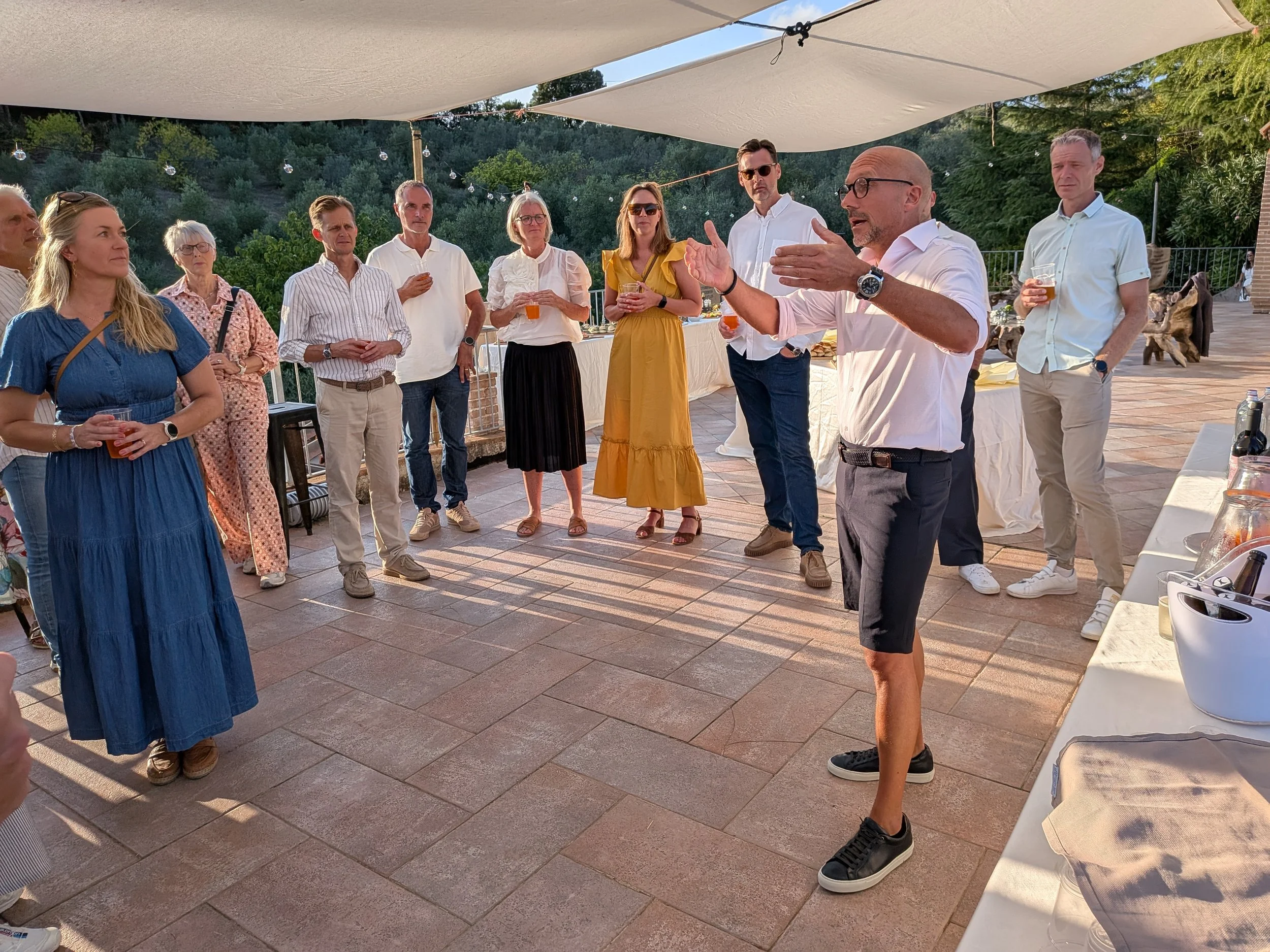 A group of people gathered outdoors at a social event, listening to a man speaking, with drinks in hand, under a shaded area during sunset.