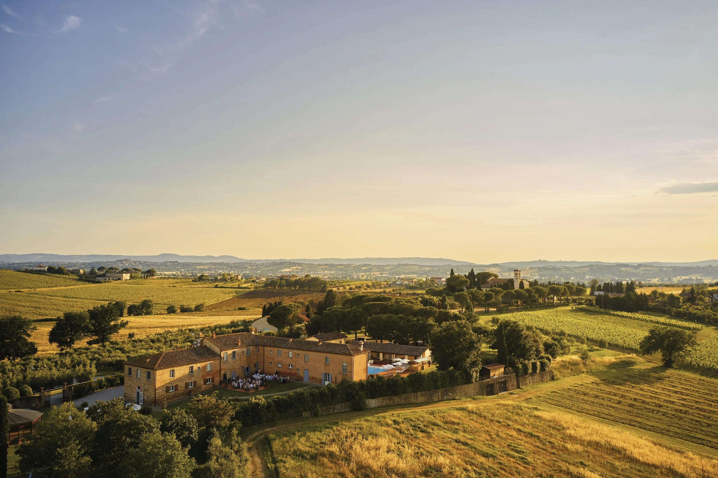 A scenic landscape view of a rustic countryside with farmhouses, trees, and fields during sunset.