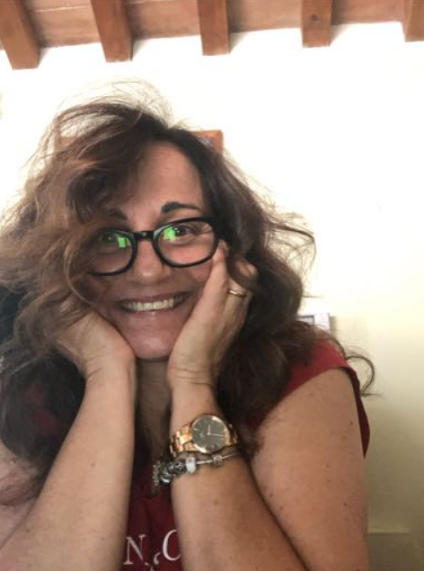 Woman with curly hair, glasses, and a red shirt, smiling and resting her face in her hands indoors.