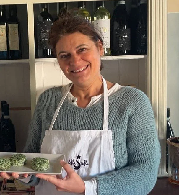 A woman smiling and holding a plate with three green leafy food items, standing in front of a shelf with wine bottles, wearing an apron over a sweater.