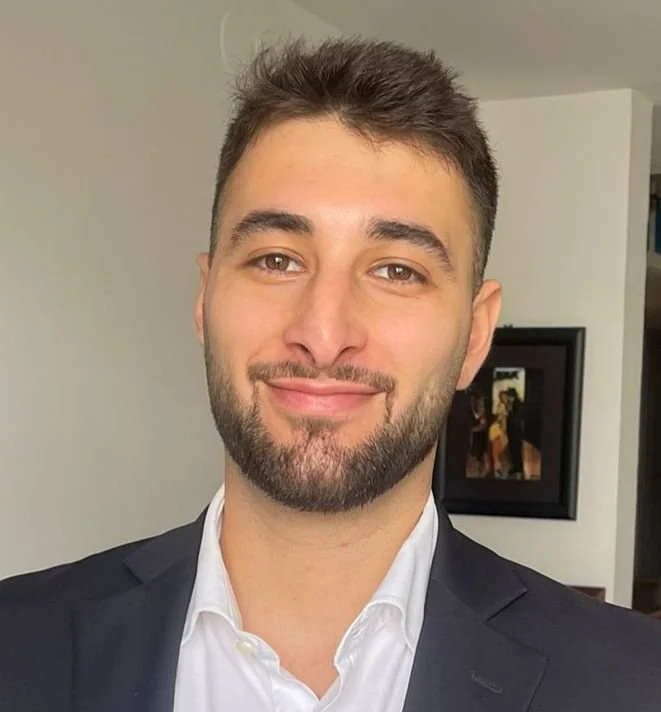 A man with dark brown hair, a trimmed beard, smiling, wearing a white shirt and dark blazer, in a room with a framed photo on the wall.