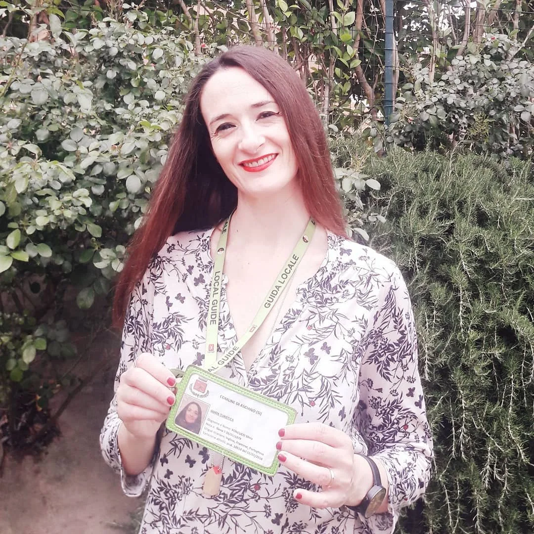 A woman with long brown hair smiling, wearing a floral patterned blouse, holding an ID card, standing outdoors among green bushes.