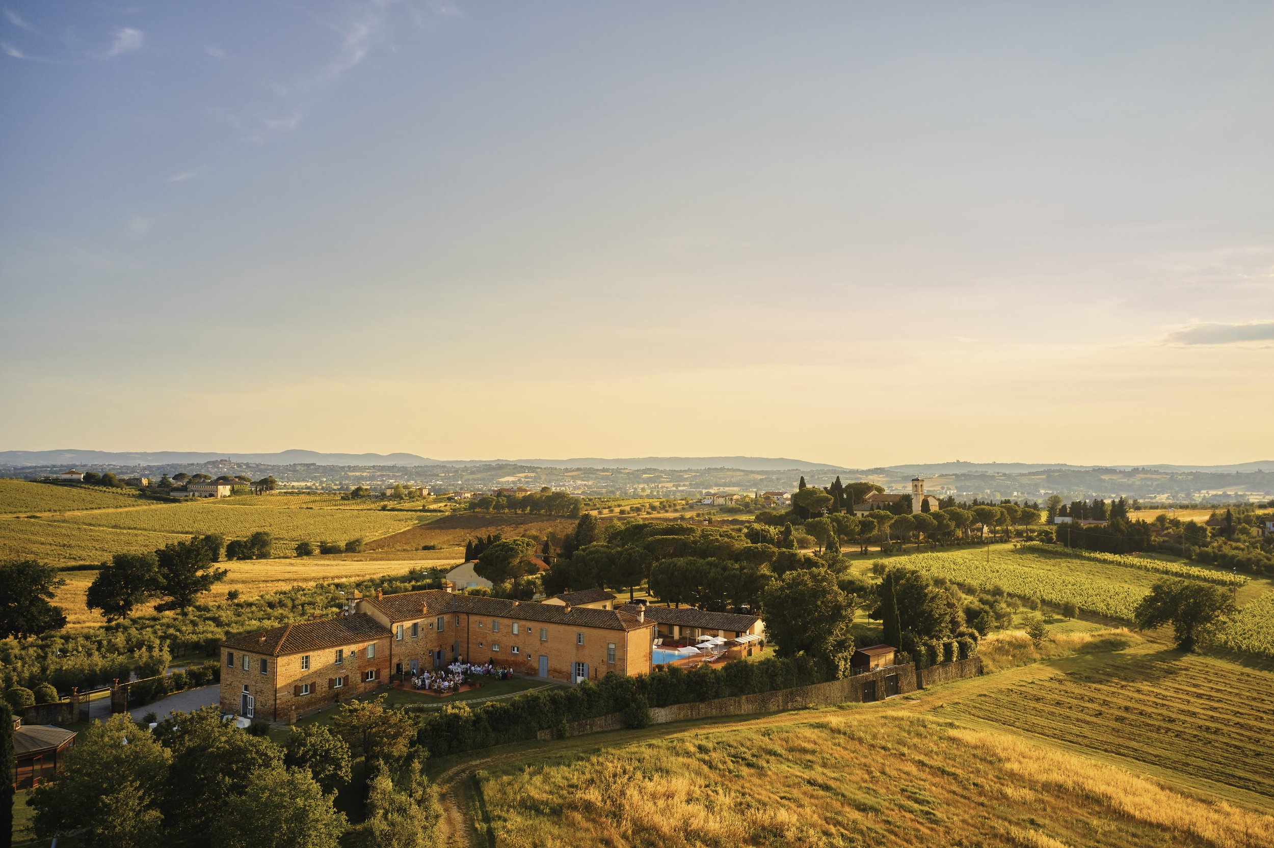 A picturesque countryside with rolling hills, fields, and a group of buildings, possibly a farmhouse or estate, surrounded by trees and open land, under a clear sky during sunset or sunrise.