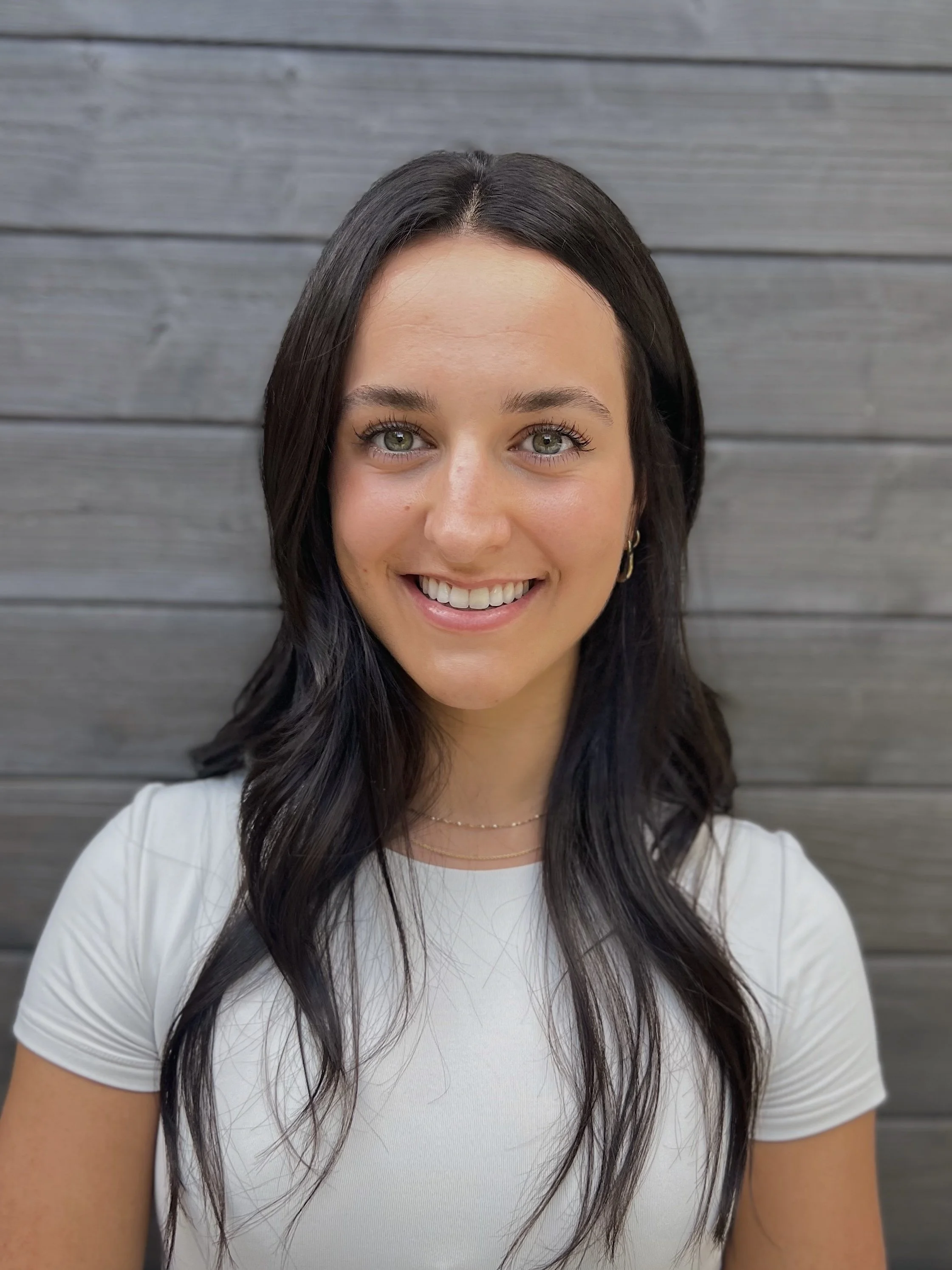 A young woman with long dark hair and light skin, smiling and wearing a white t-shirt, standing in front of a gray wooden wall.