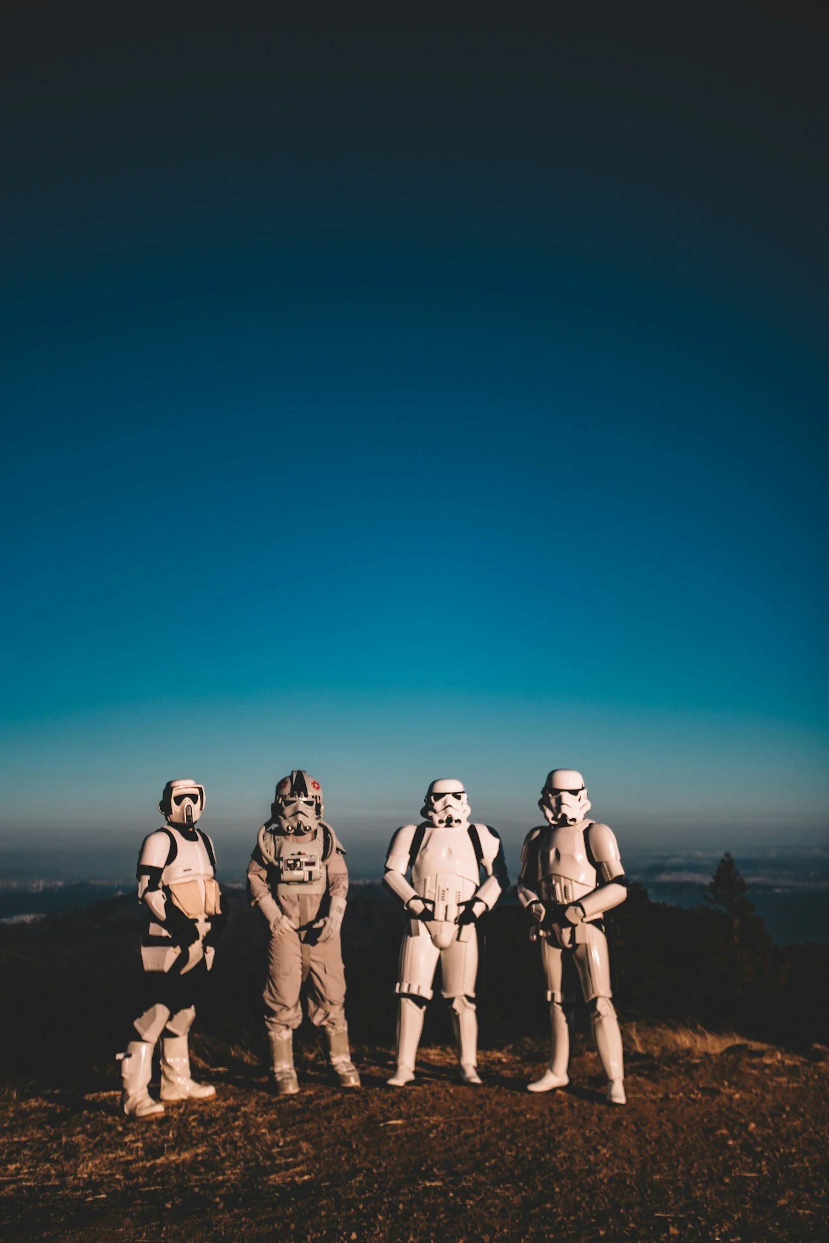 Four people dressed as Star Wars stormtroopers and a clone trooper standing on a hilltop during dusk, with a cityscape in the distance and a clear, darkening sky above.