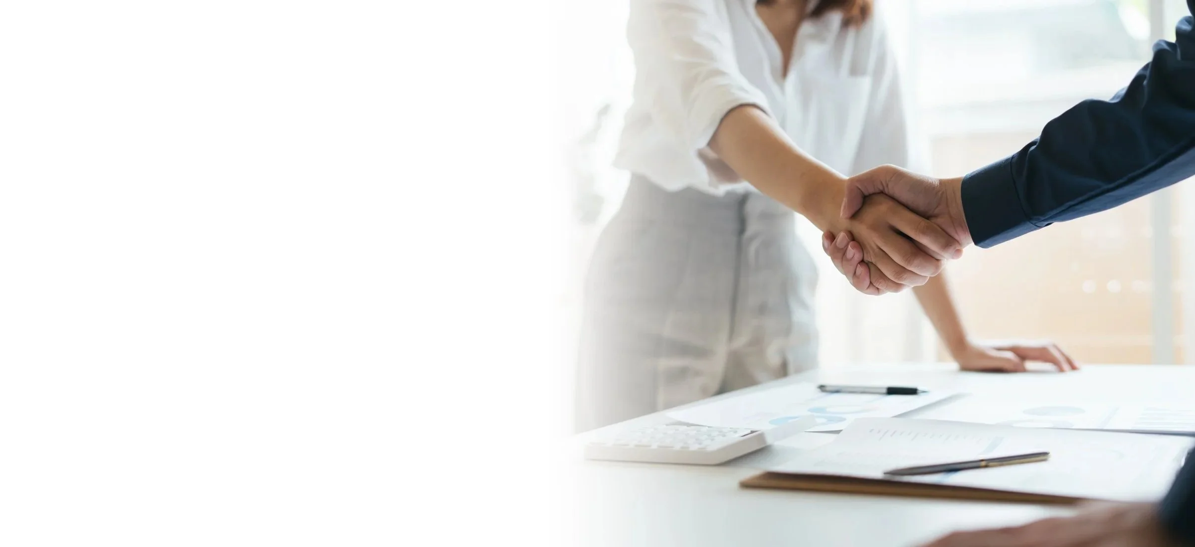 Two people in business suits shaking hands over a table with documents and charts.