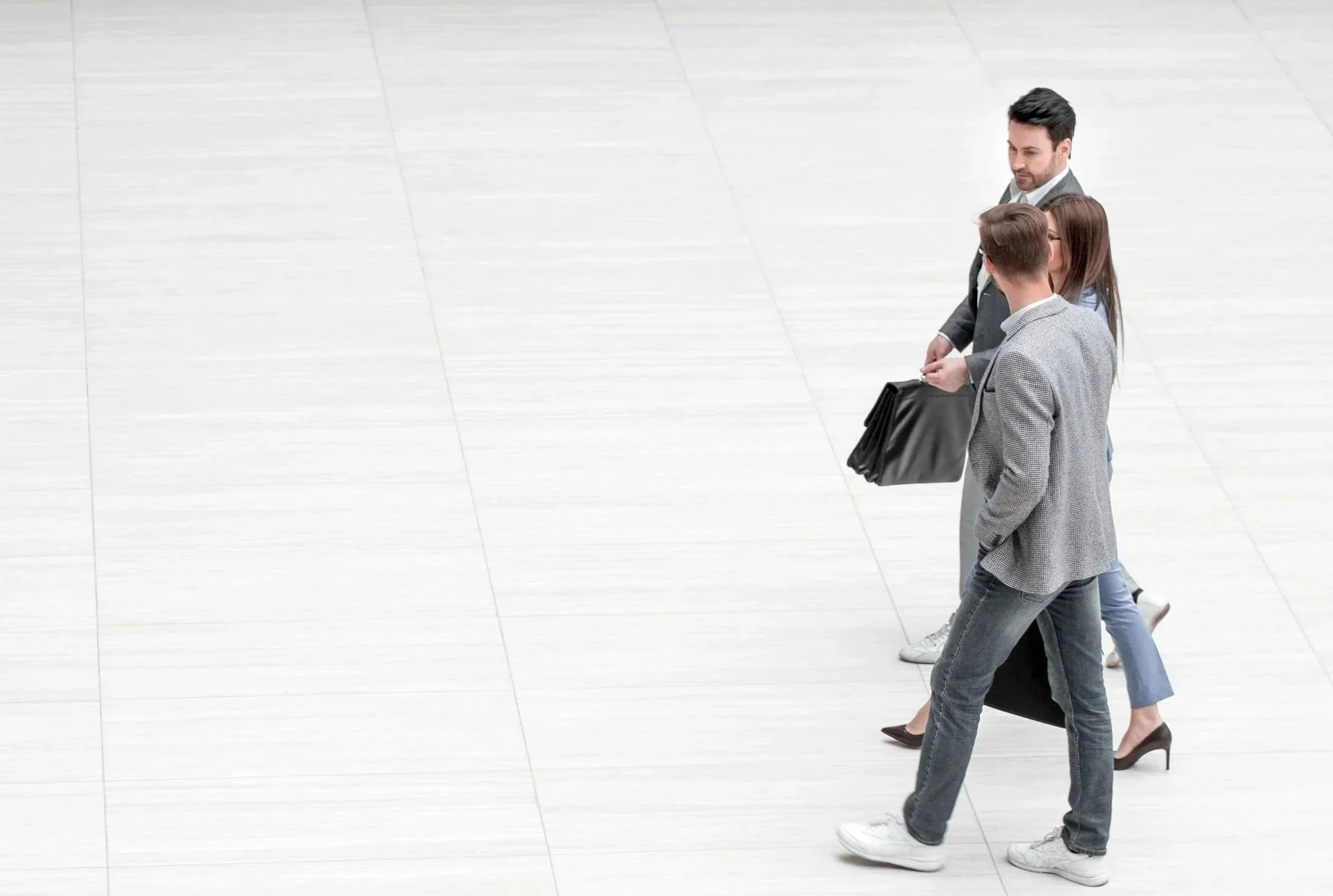 Three business people walking on a light-colored tiled floor, engaged in conversation, with the men dressed in suits and the women in high heels.