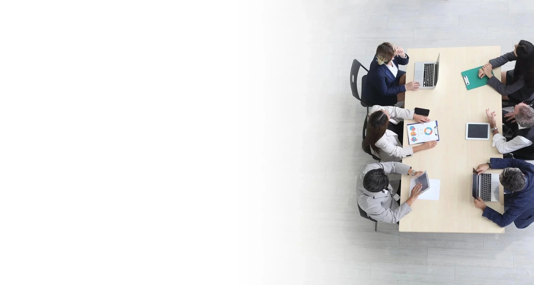 Eight businesspeople sitting around a rectangular conference table engaged in discussion with tablets and laptops.