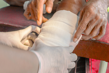 Someone is bandaging an elderly person's arm with a white cloth, while another person wearing gloves assists with the process.