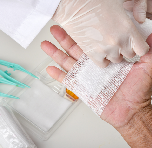A healthcare worker wearing gloves performing a procedure on a patient's wrist with medical tools and supplies nearby.