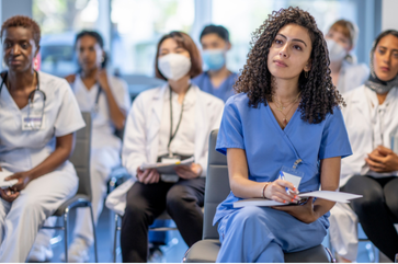 Group of diverse healthcare students in a classroom, listening attentively with some taking notes, wearing medical scrubs and masks.