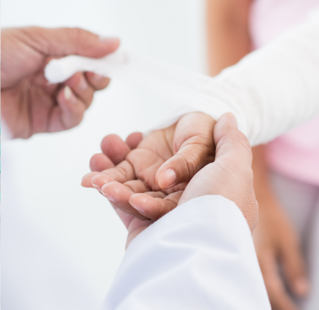 A person in a white medical coat bandages the arm of a patient, who is reclining, with the patient's hand resting on a healthcare worker's palm.