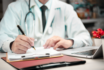 Close-up of a doctor writing in a clipboard with a pen, wearing a white coat and stethoscope, with a laptop and red flowers on the desk.