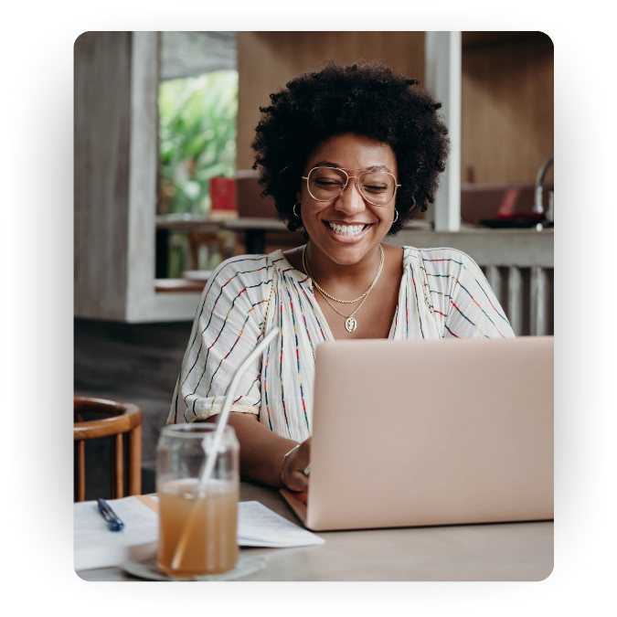A smiling woman with curly hair and glasses working on a laptop at a cafe, with a glass of iced coffee and a notebook on the table.