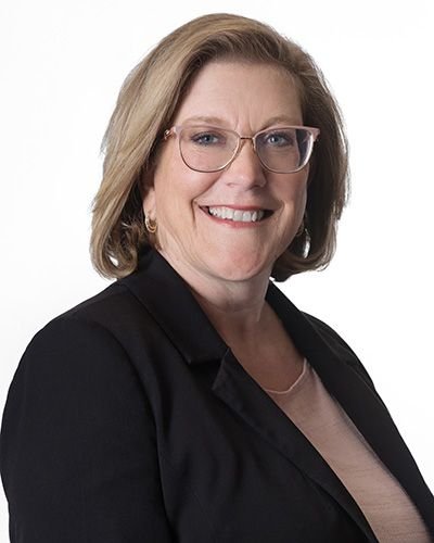 A smiling woman with glasses, shoulder-length blonde hair, wearing a black blazer and a light-colored top against a white background.