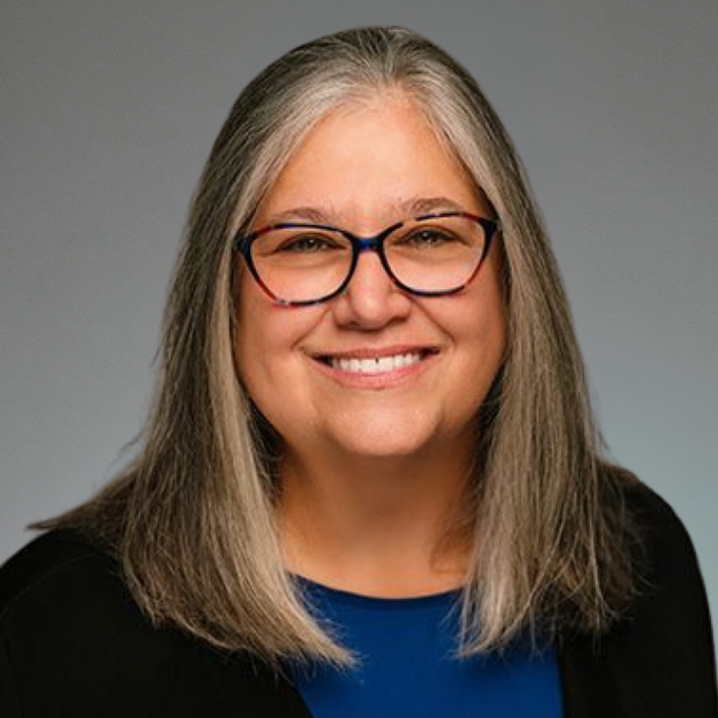 Headshot of a smiling woman with gray hair, glasses, wearing a black blazer and blue top, against a neutral gray background.