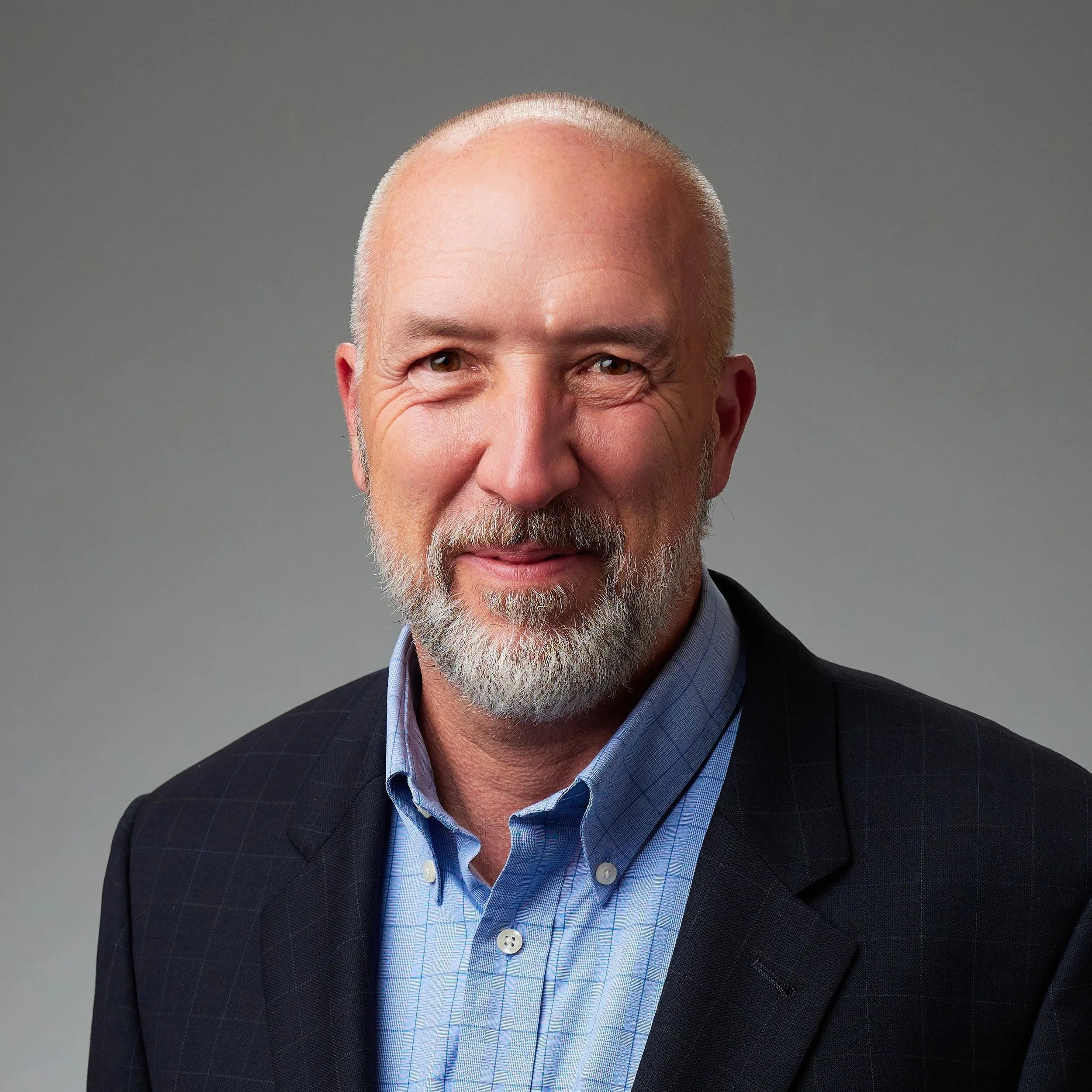 Headshot of an older man with a beard, wearing a dark suit jacket and a blue collared shirt, standing against a plain gray background.