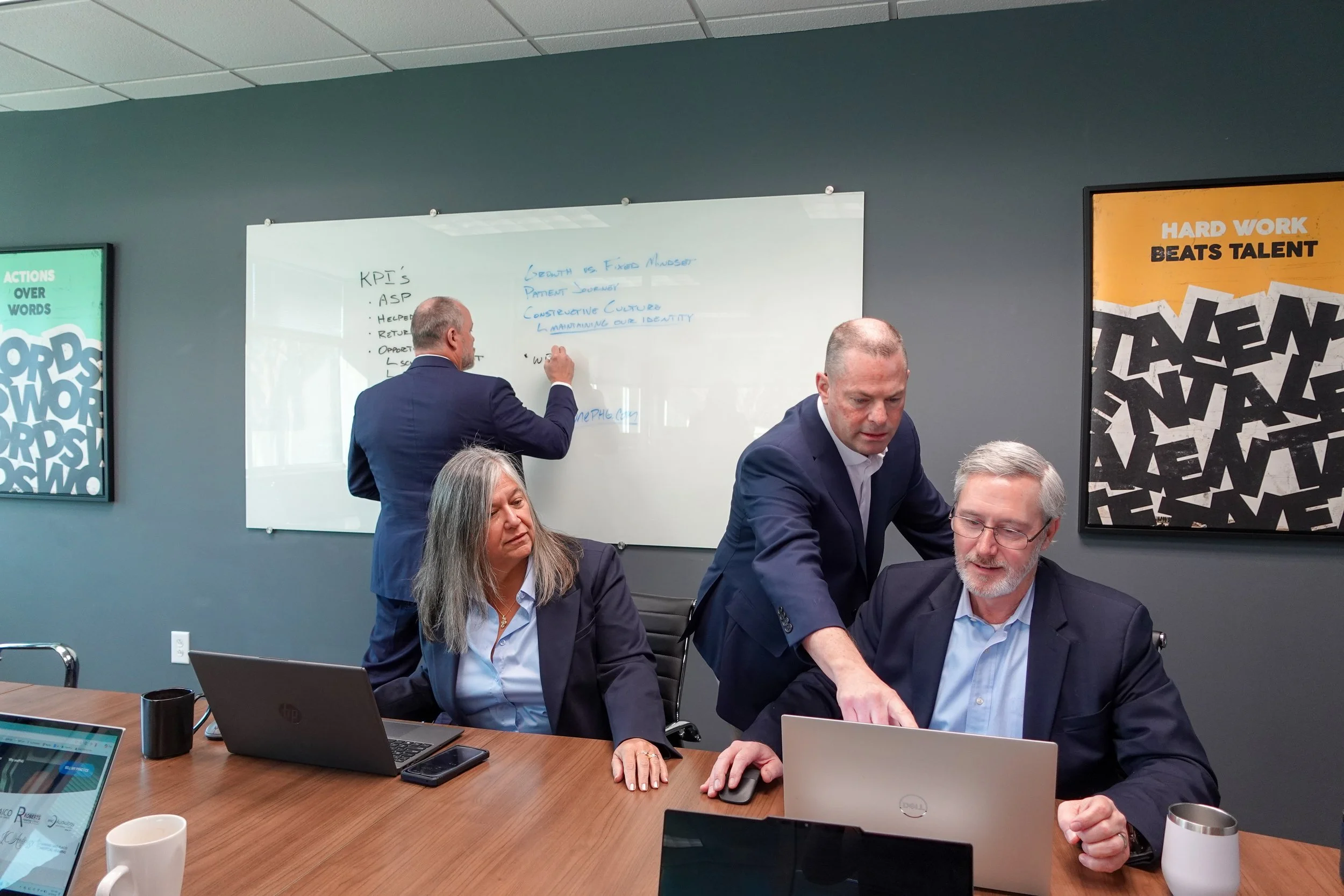 Business meeting in a conference room with people working on laptops and discussing. Whiteboard with handwritten notes, and wall posters with motivational phrases.