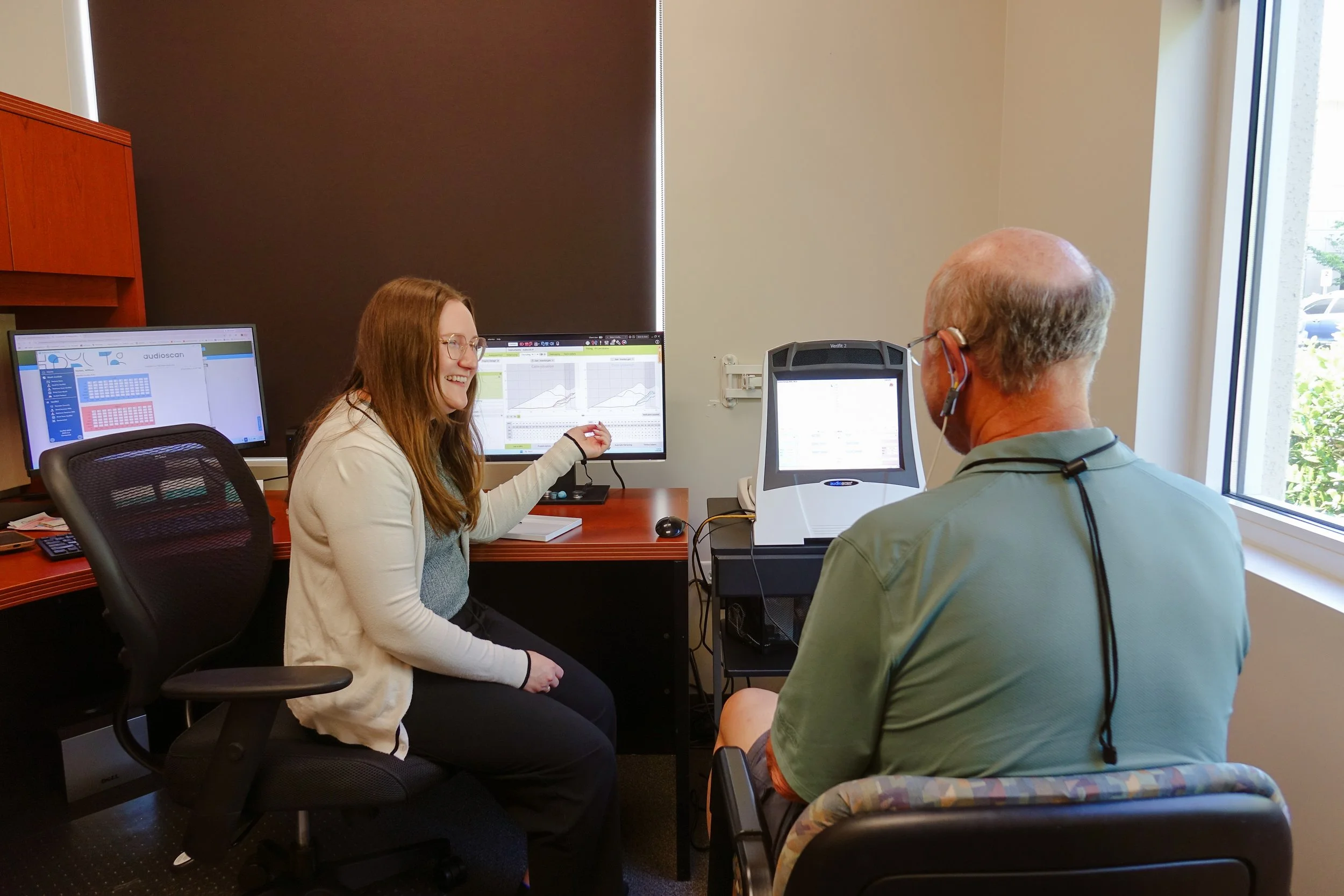 A woman with long brown hair and glasses is smiling and sitting at a desk with two computer monitors displaying graphs and charts. She is talking to an older man with glasses, who is sitting across from her with his back to the camera, near a window with blinds. The room has beige walls and office furniture.