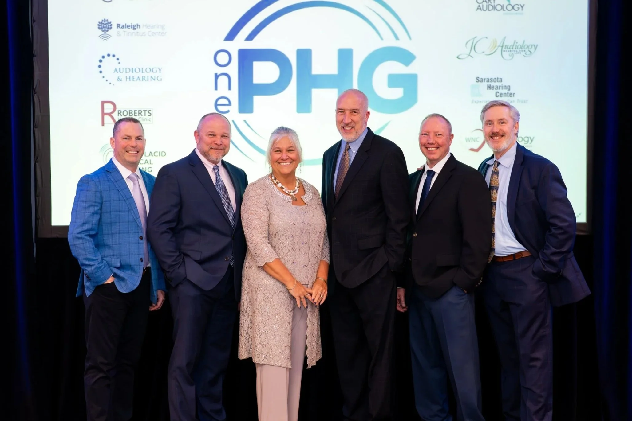 Group of seven professionally dressed individuals standing together in front of a presentation screen. The screen displays the logo for the 'One PHG' event and logos of various hearing and audiology centers.
