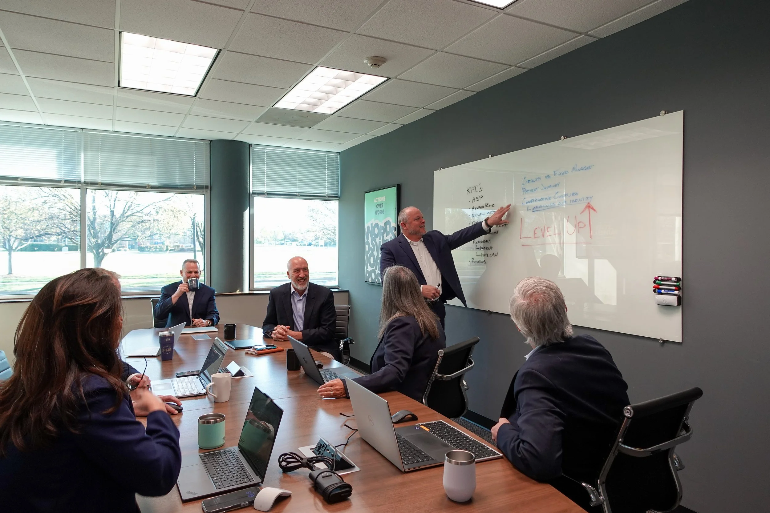A business meeting taking place in a conference room with several people seated around a rectangular table. One man is standing near a whiteboard, gesturing as he speaks. Others are listening, some with laptops open. There are large windows with blinds allowing natural light, and the room has a professional atmosphere.