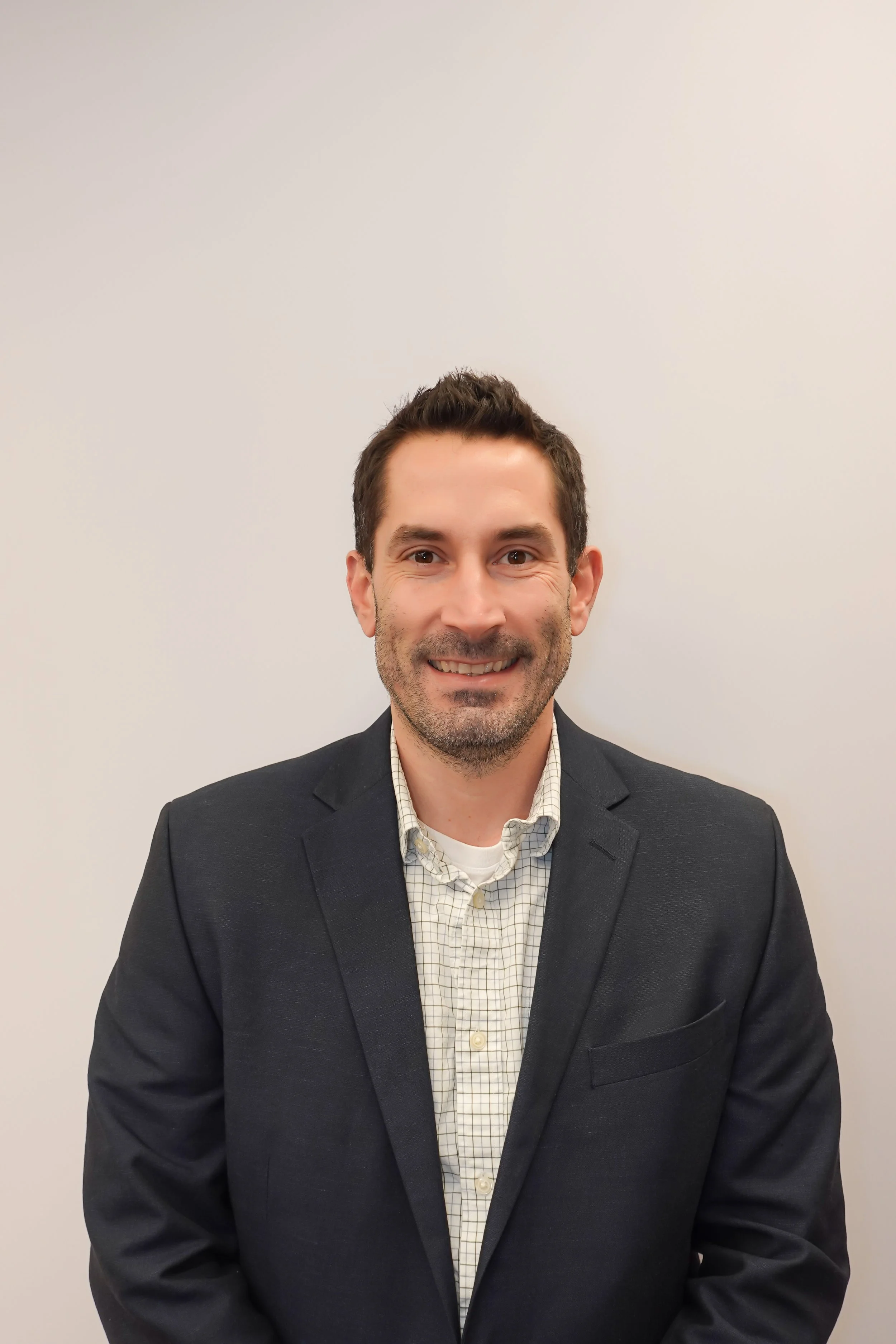 A man wearing a dark suit and a checked dress shirt, smiling and standing against a plain, light-colored wall.