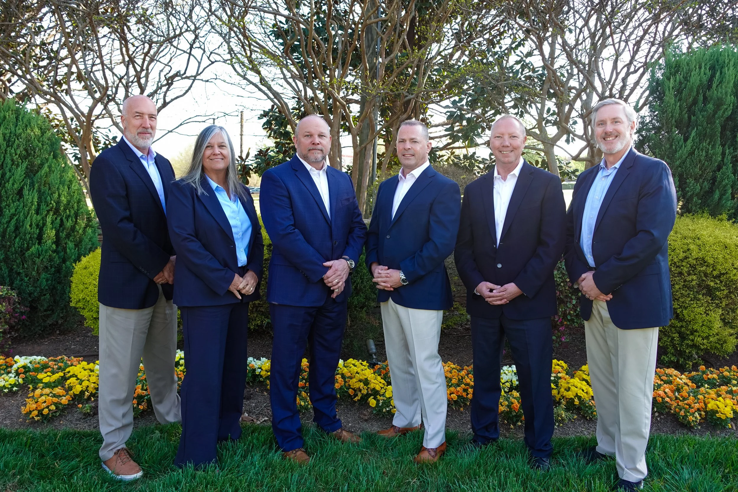 Seven adults standing outdoors in front of trees and colorful flowers, dressed in business casual attire, posing for a group photo.