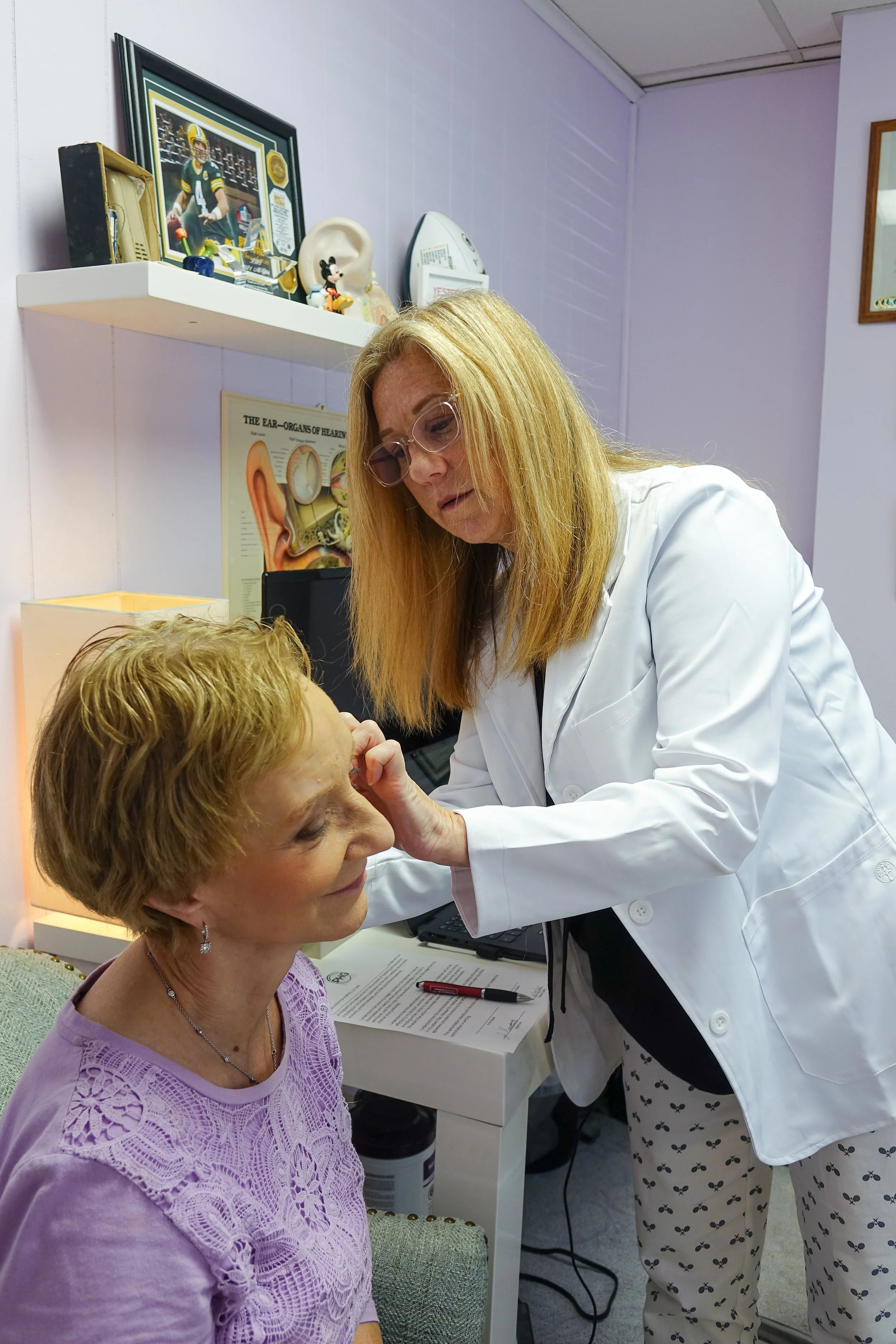 A woman with red hair and glasses is examining a woman with short, curly, blonde hair and earrings, who is seated, in a medical or clinical setting.