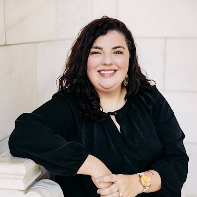 A woman with dark, curly hair smiling and leaning on a white surface, wearing a black top and a gold watch.
