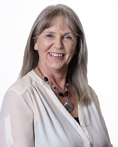 A smiling woman with gray hair wearing a white blouse and a black necklace with a pendant, against a plain white background.