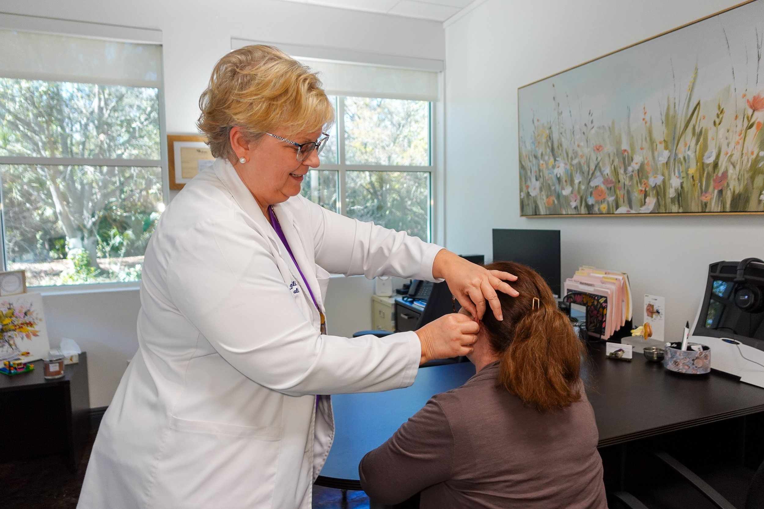 A healthcare professional in a white coat examining a patient's head in an office with large windows and floral artwork.