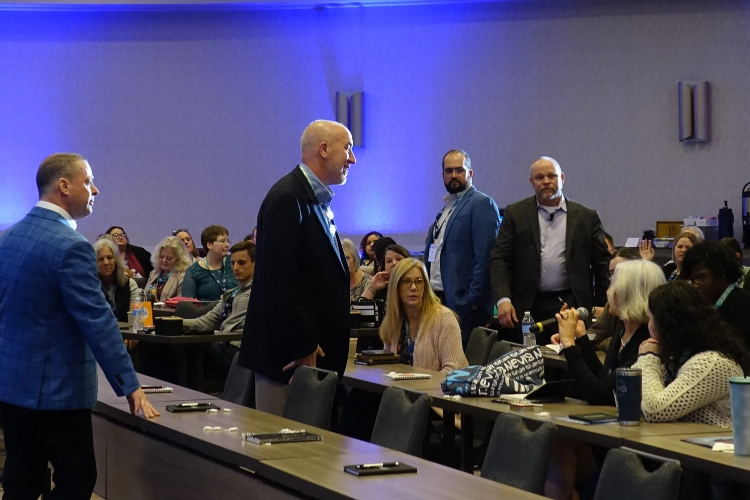 Conference room with people seated at tables, some standing and engaging in conversation, with a plain wall and blue lighting in the background.