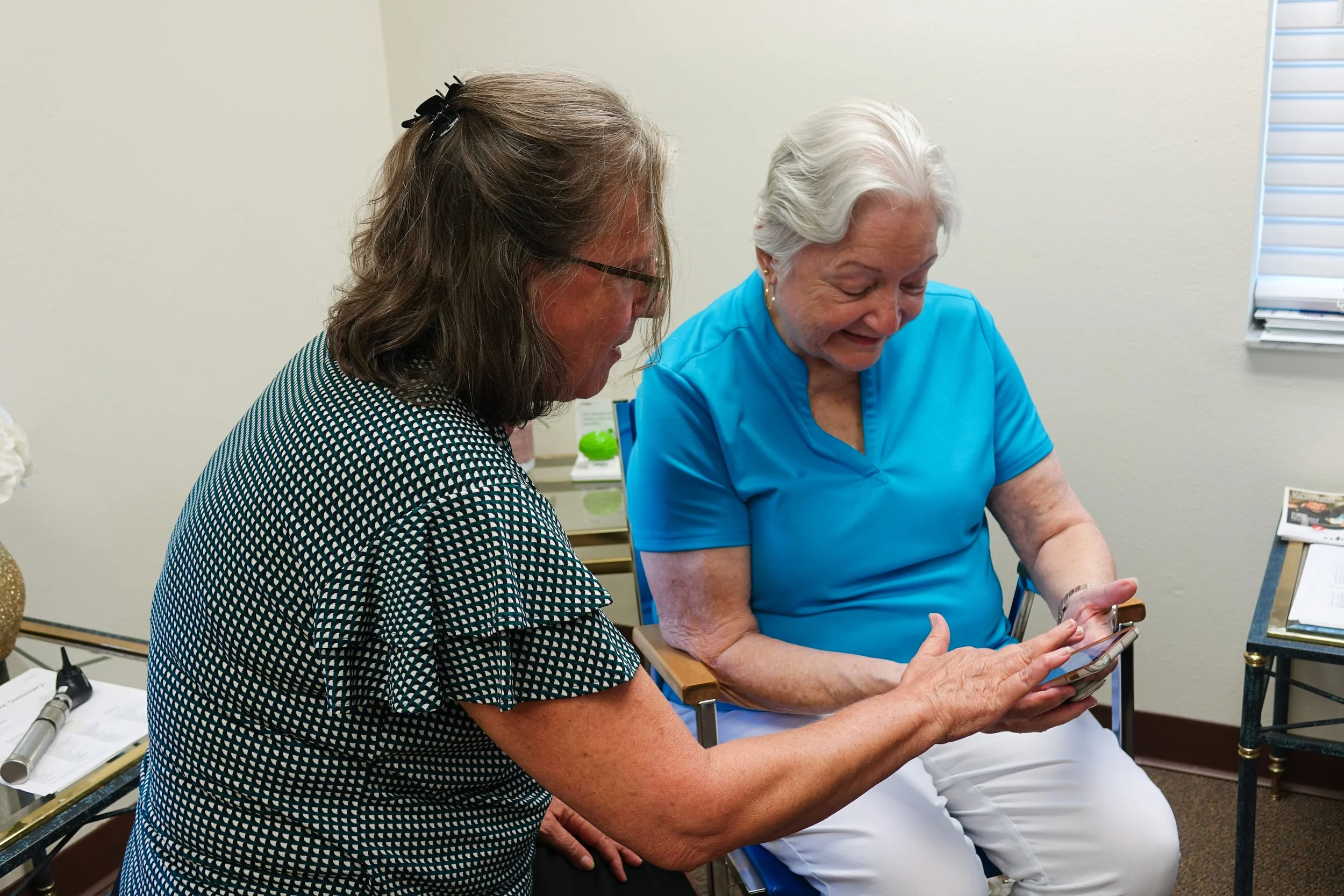 Two women, one middle-aged with brown hair and glasses, and the other elderly with gray hair, are sitting and looking at a mobile phone together in a room with medical equipment and documents.