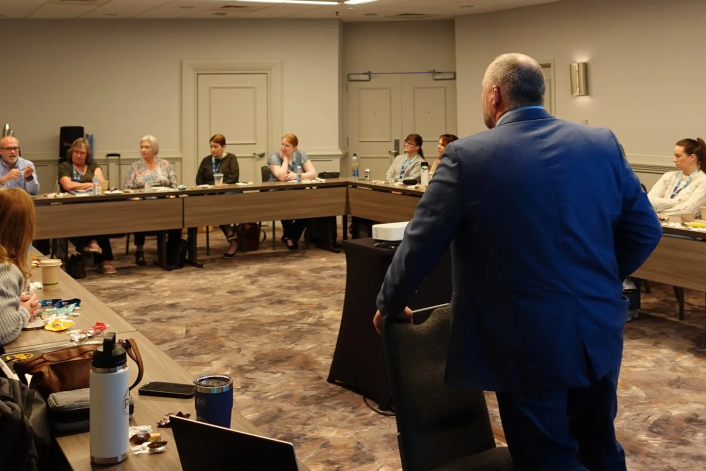 A man in a blue suit stands at a conference table in a meeting room with several women seated around the table, listening to him.