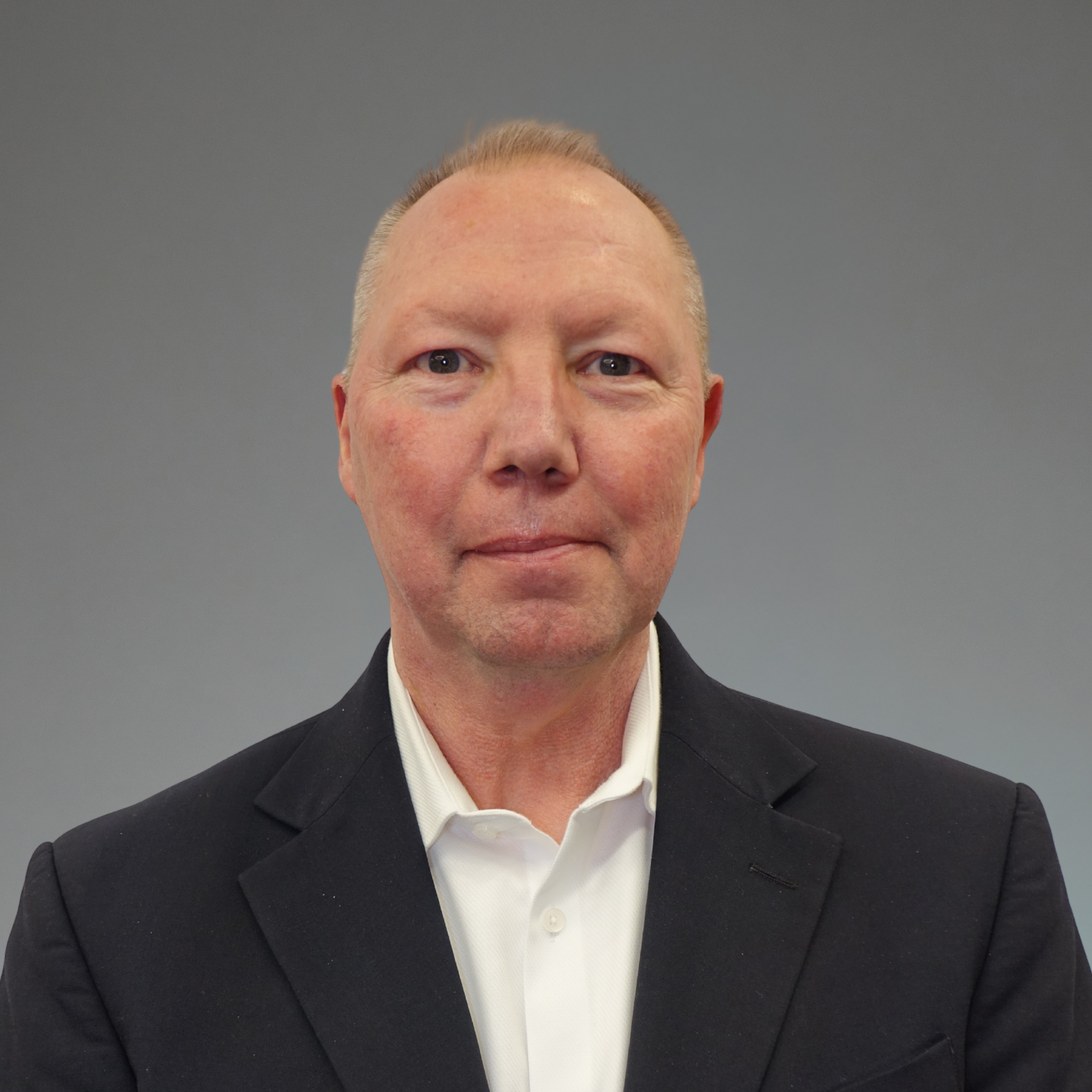Headshot of a middle-aged man with light skin, short light brown hair, wearing a black suit jacket and white shirt against a plain gray background.
