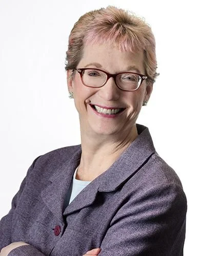 Portrait of an older woman with short light hair, wearing glasses, a gray blazer, and a white shirt, smiling against a plain background.