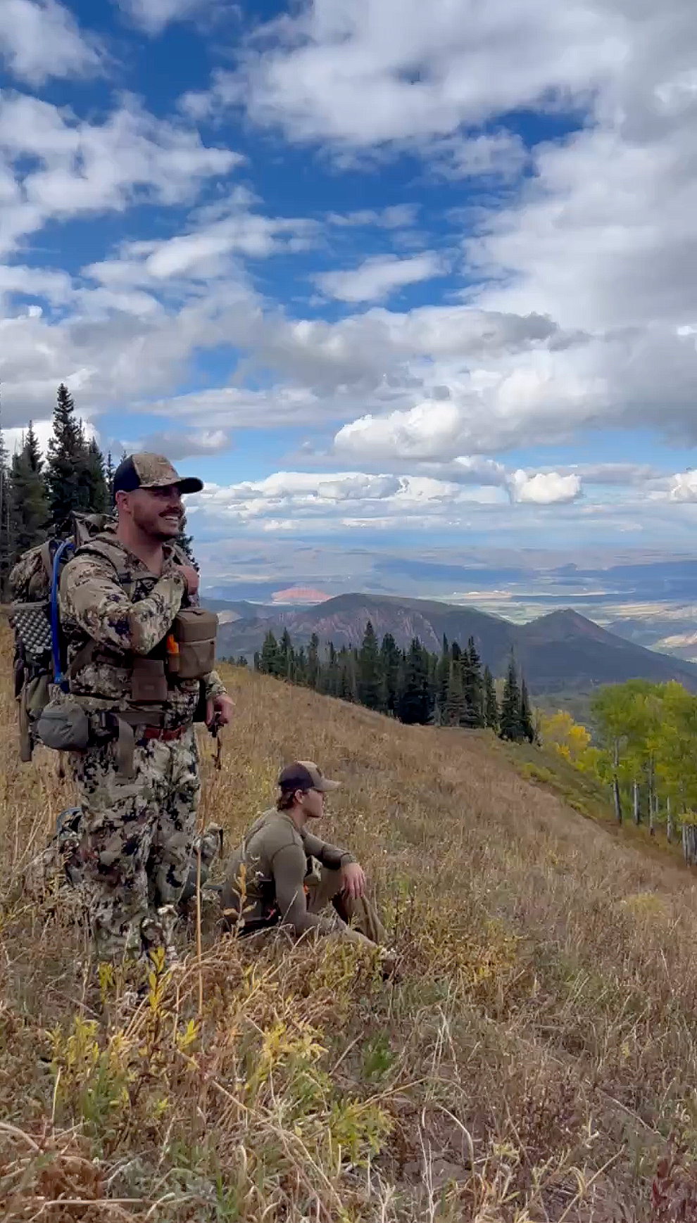 Two hunters resting on a grassy hillside overlooking a valley with mountains in the distance under a partly cloudy blue sky.