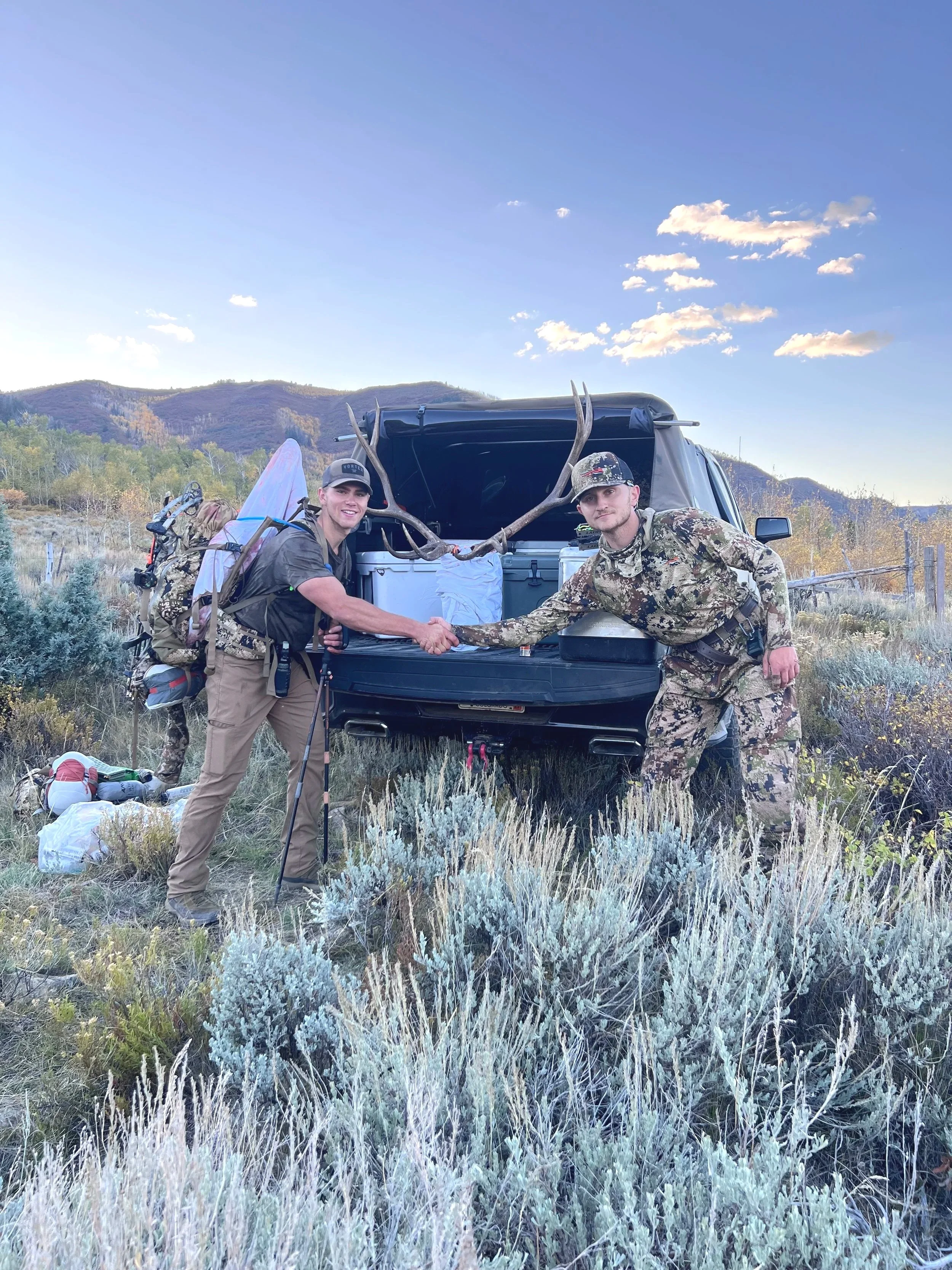 Two hunters shaking hands in front of a black vehicle with large antlers on the tailgate, in a rural outdoor setting with hills and shrubbery in the background.