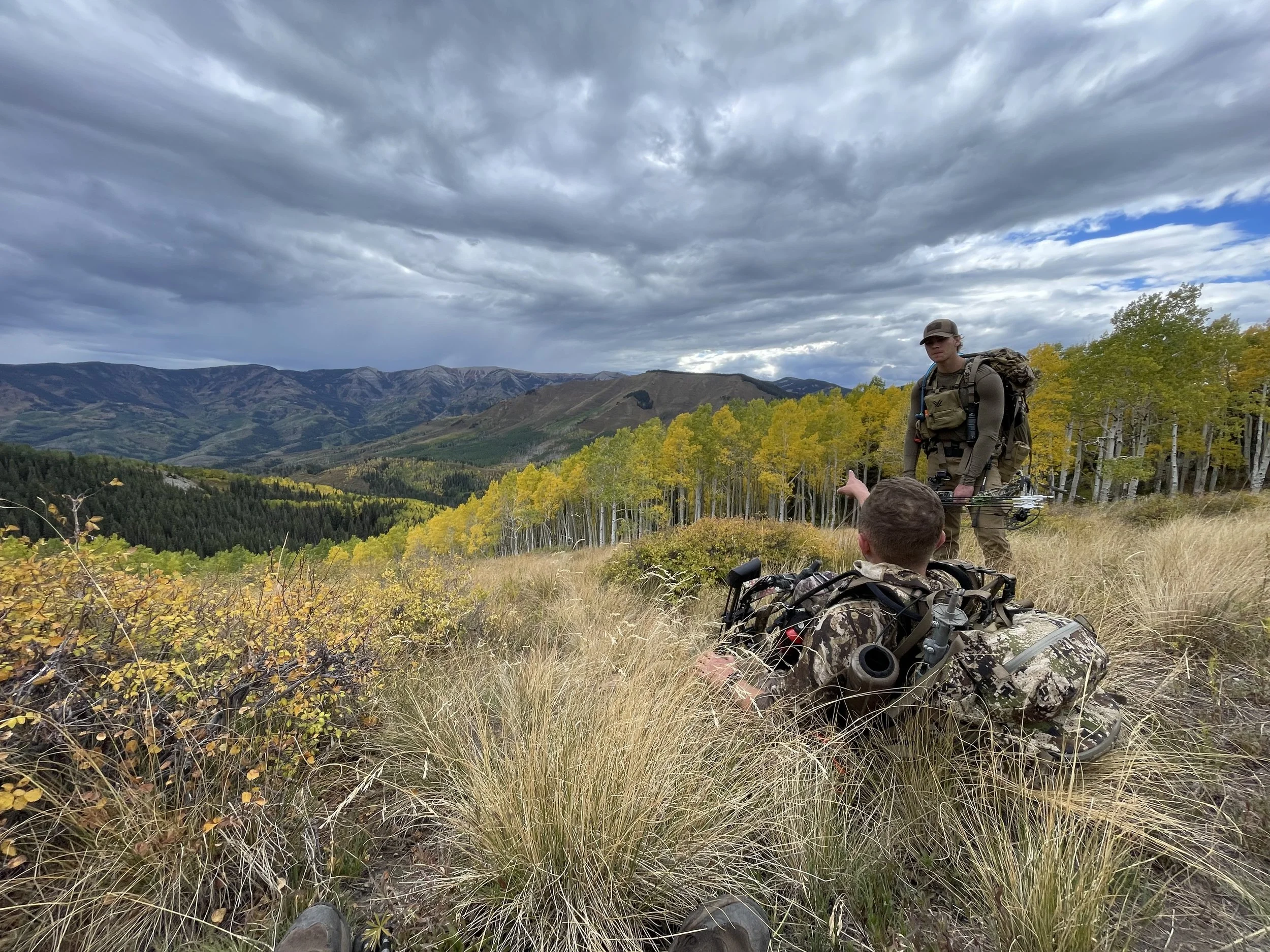 Two hunters resting on a grassy hill in a mountain landscape, one sitting on the ground and gesturing, the other standing with binoculars, surrounded by trees and mountains under a cloudy sky.