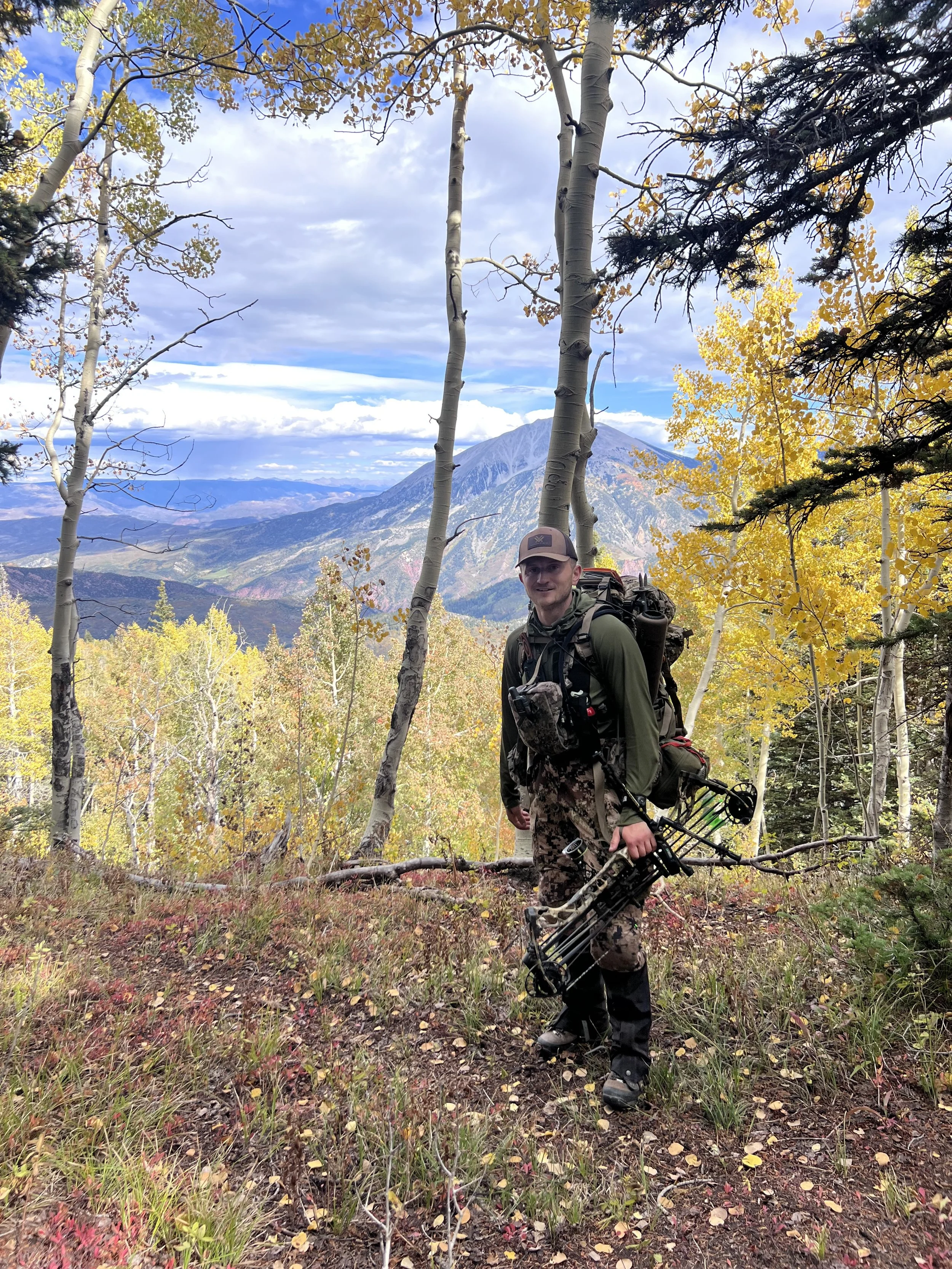 Hunter in camouflage lifting bow in a forest with fall-colored trees and mountains in the background.