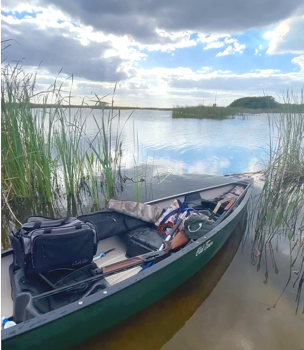 A green canoe filled with gear resting in shallow water near tall grass at the water's edge, with a partly cloudy sky reflected in the water and a small island or landmass in the distance.
