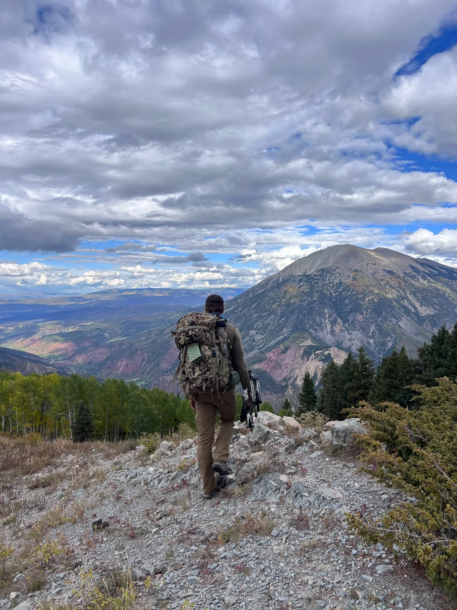 Hunter with a backpack hiking on a rocky trail in a mountainous landscape with trees and a view of mountain peaks under a partly cloudy sky.