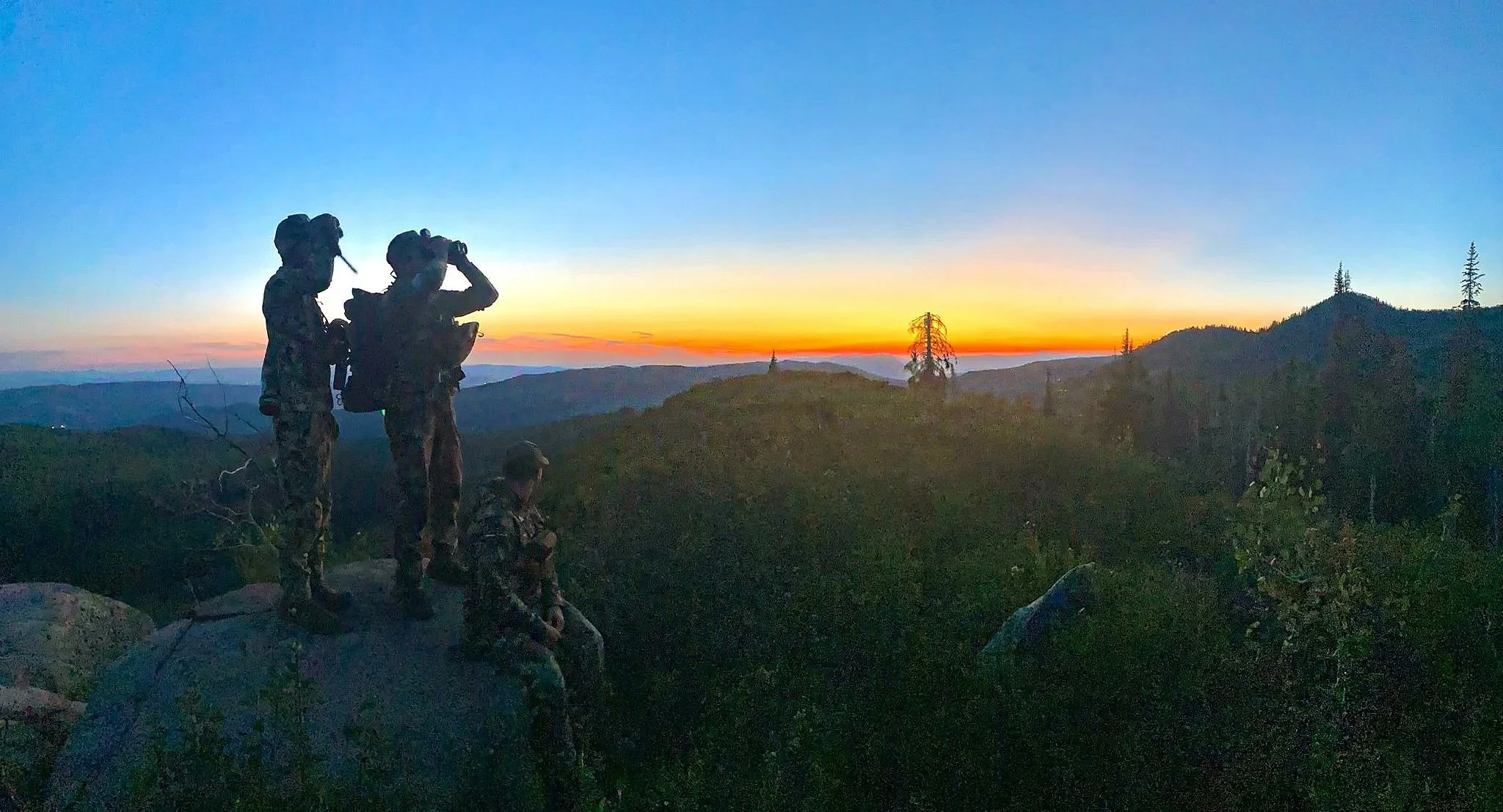 Four hunters in camouflage uniforms watching a sunset on a mountain top, with one using binoculars and two sitting on rocks.