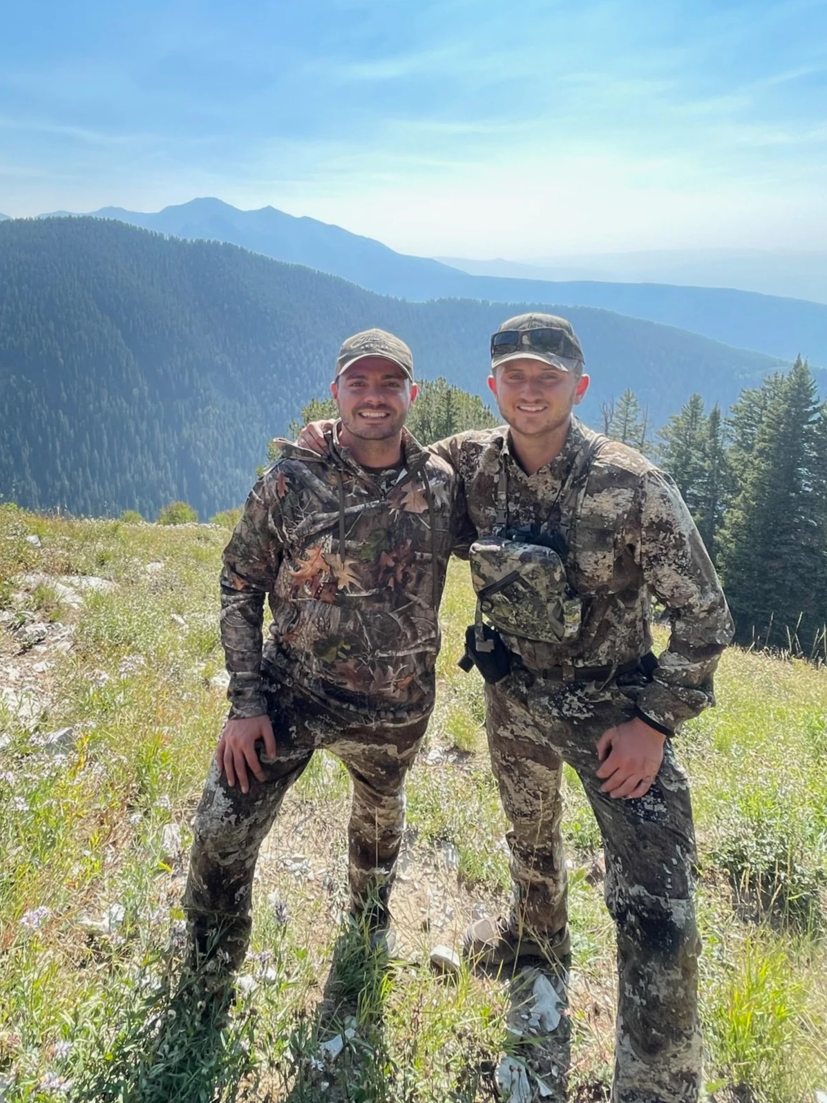 Two hunters standing outdoors in a mountainous landscape, smiling and posing with their arms around each other.