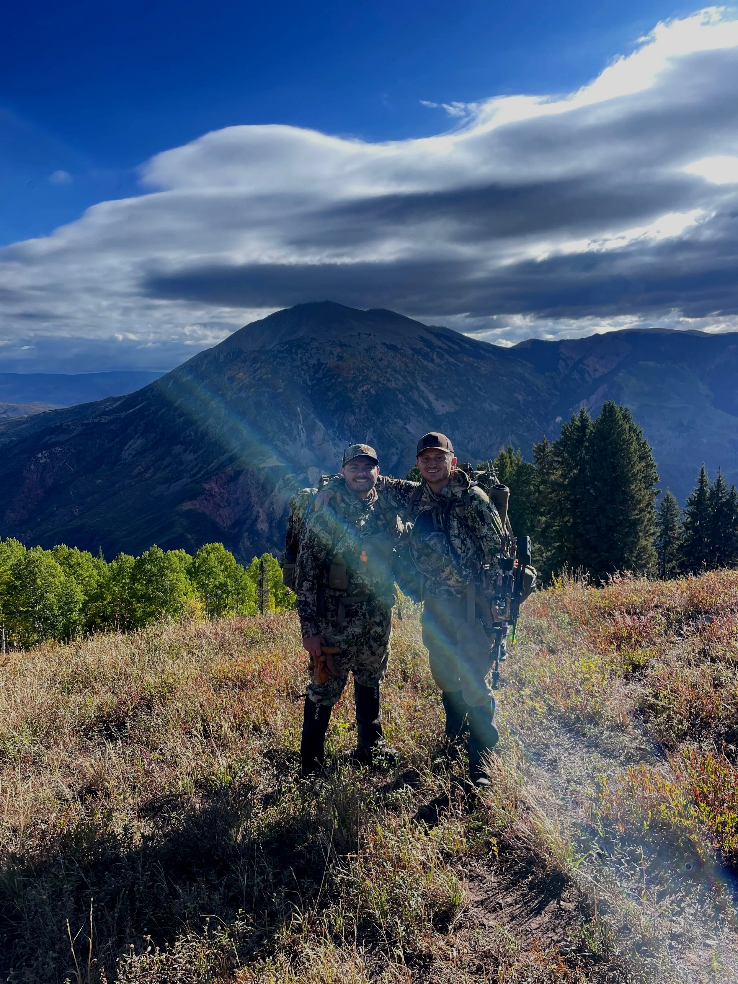 Two hunters in camouflage gear standing on a grassy mountain slope with trees, a large mountain, and cloudy sky in the background.