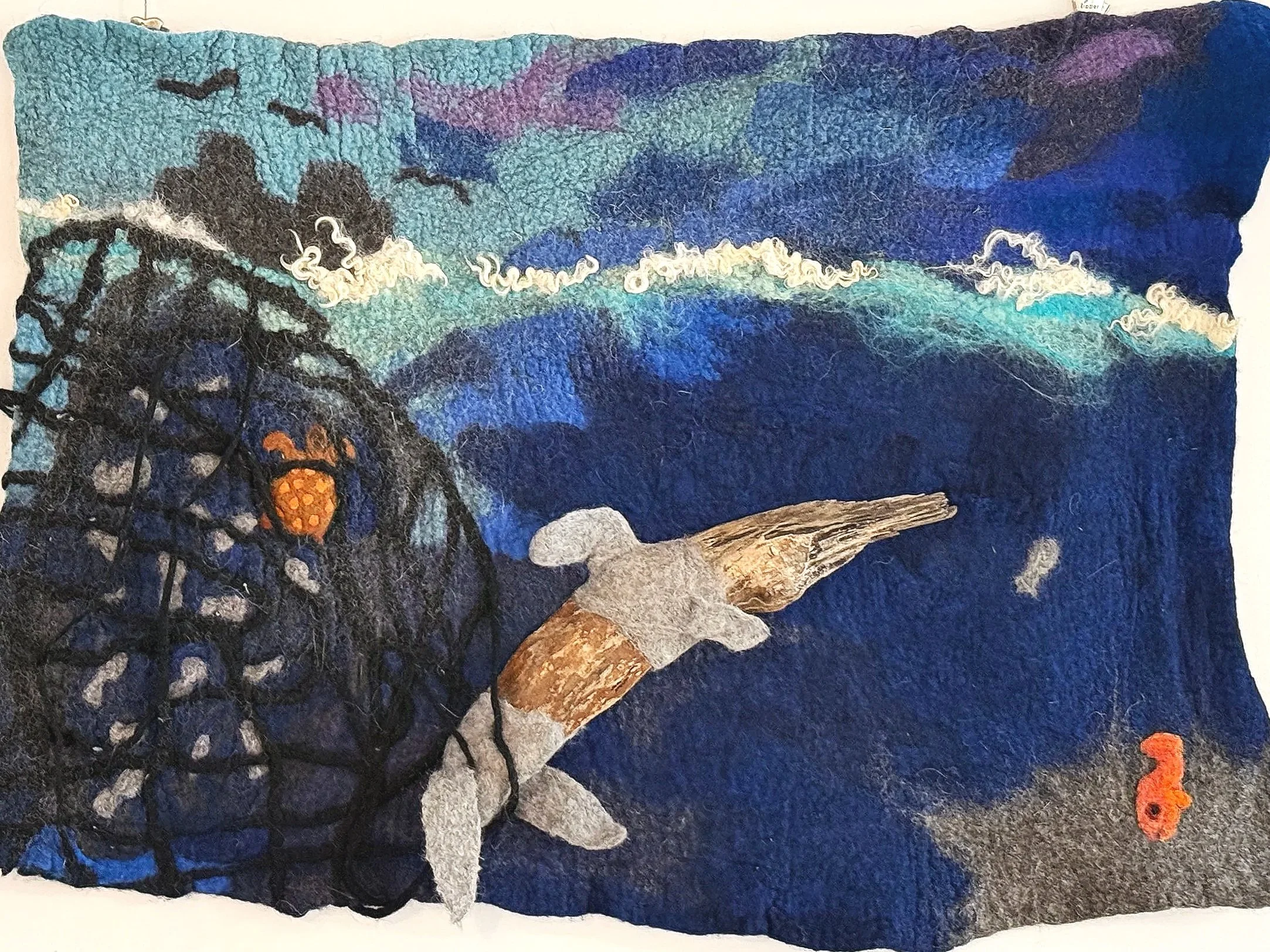 Empty Waters – 92 × 68 cm
Material: Wolle, Filz, ein Holzfundstück (Delfin), Netz, Wollfasern

Das Netz aus dem ersten Bild wächst weiter und wird zum Symbol der Überfischung. Links hängt Beifang fest: eine orange Schildkröte und ein Delfin aus einem